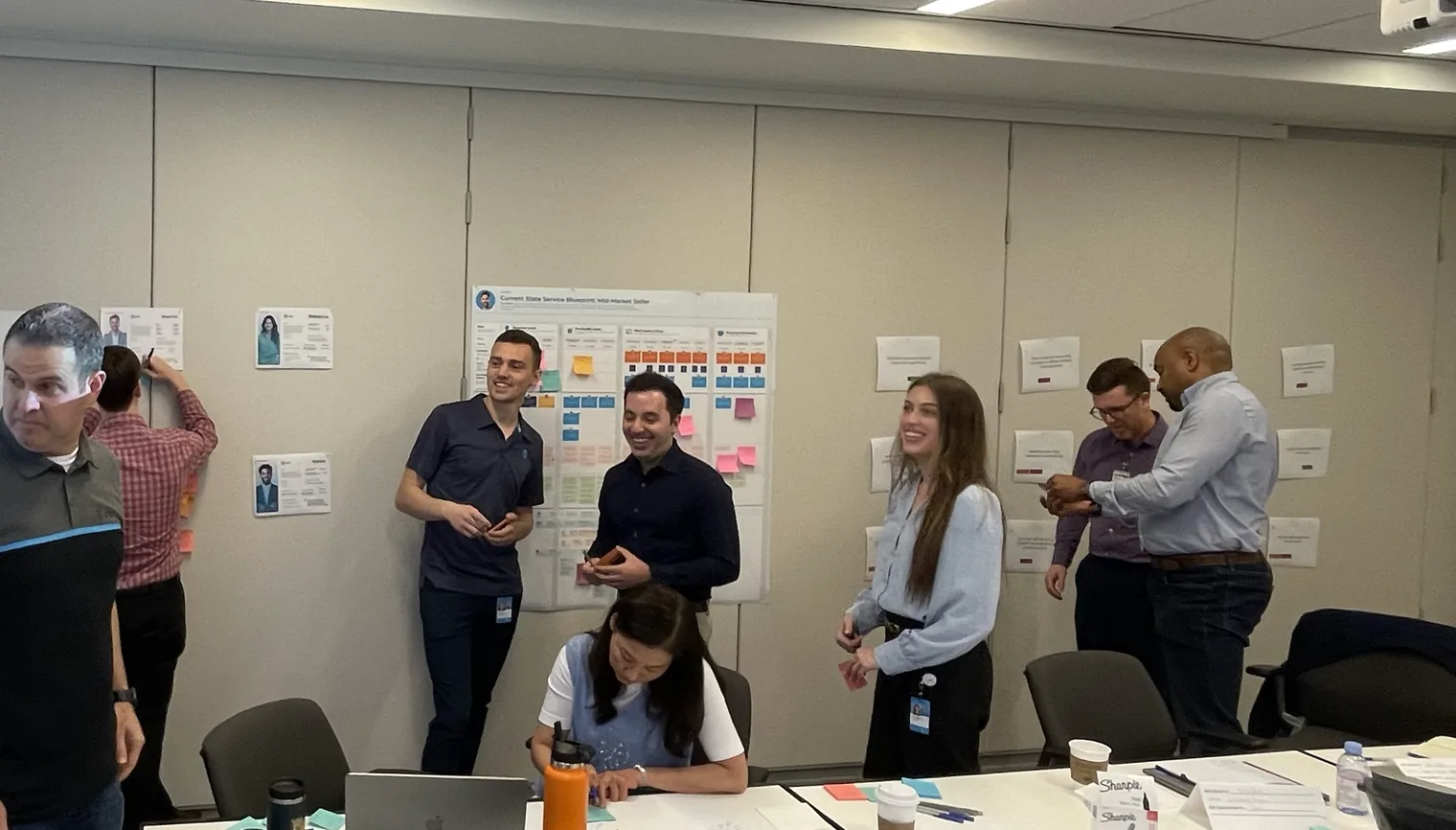 A Fyve Labs working session. Participants stand at a wall-sized service blueprint covered in sticky notes, mid-discussion.